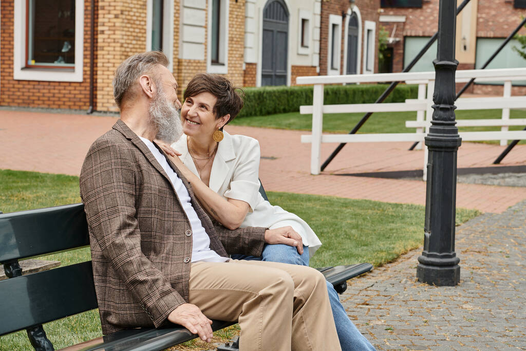 happy elderly woman looking at bearded man, romance, husband and wife sitting on bench, urban - Photo, Image