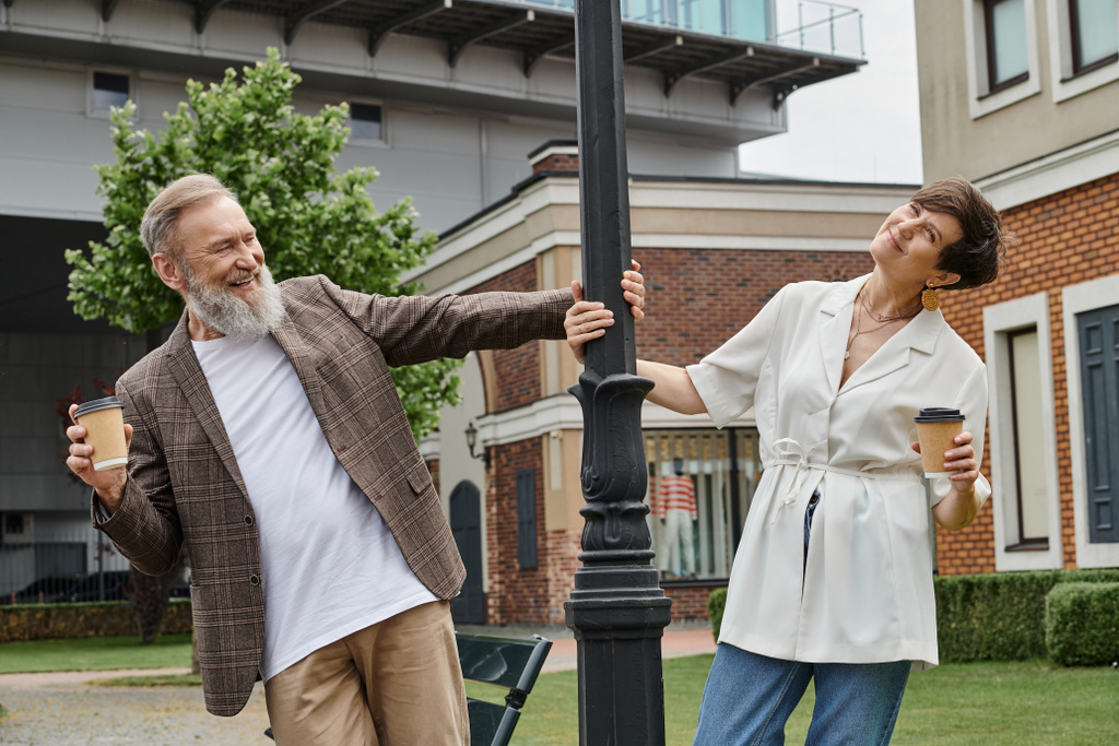 feliz pareja de ancianos sosteniendo café para llevar, de pie cerca de la lámpara de calle, hombre y mujer, ancianos, romance - Foto, Imagen