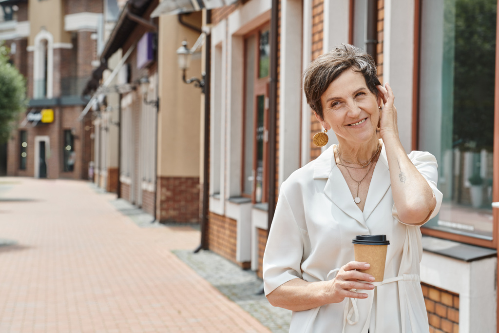 щаслива старша жінка тримає паперову чашку з кавою, коригуючи коротке волосся, міський фон, посміхається - Фото, зображення