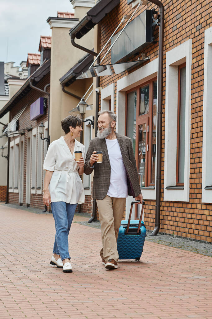 feliz pareja de ancianos, hombre y mujer caminando con café para llevar y equipaje en la calle, estilo de vida urbano - Foto, imagen
