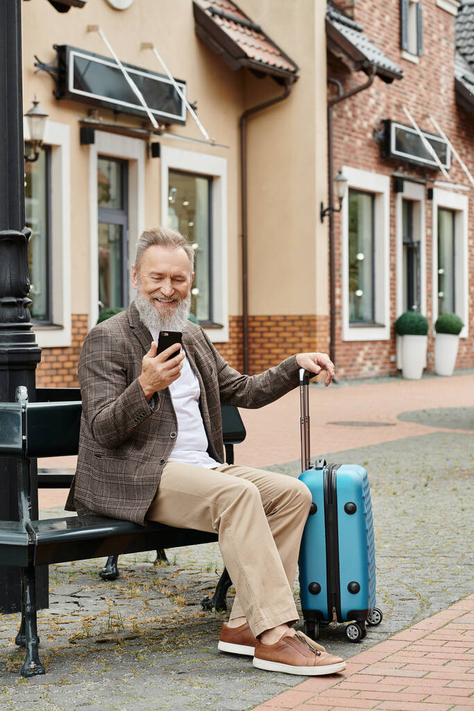 derűs idős férfi használ okostelefon és ül padon közelében poggyász, használata modul, kor tech - Fotó, kép