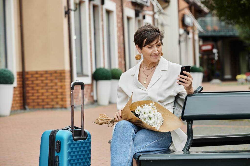 elderly woman with short hair using smartphone, holding bouquet, sitting on bench near luggage - Photo, Image