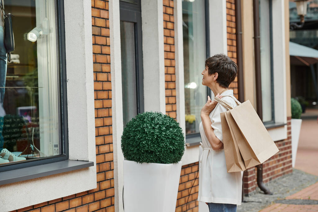 mulher sênior com cabelo curto segurando sacos de compras e olhando para a janela do shopping ao ar livre, tomada - Foto, Imagem