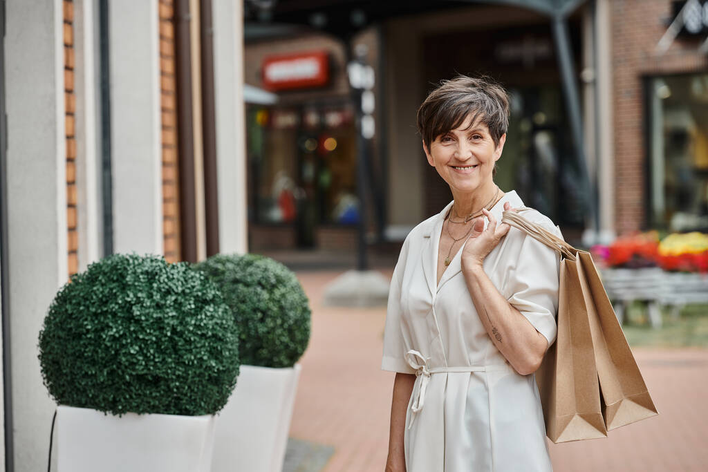 gelukkig senior vrouw met kort haar houden boodschappentassen en kijken naar camera, outdoor winkelcentrum, stopcontact - Foto, afbeelding