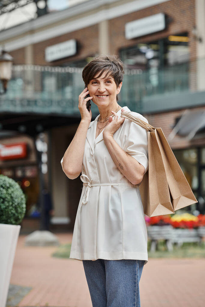 glückliche ältere Frau, die mit dem Smartphone spricht, Einkaufstüten hält und in der Nähe eines Einkaufszentrums steht - Foto, Bild
