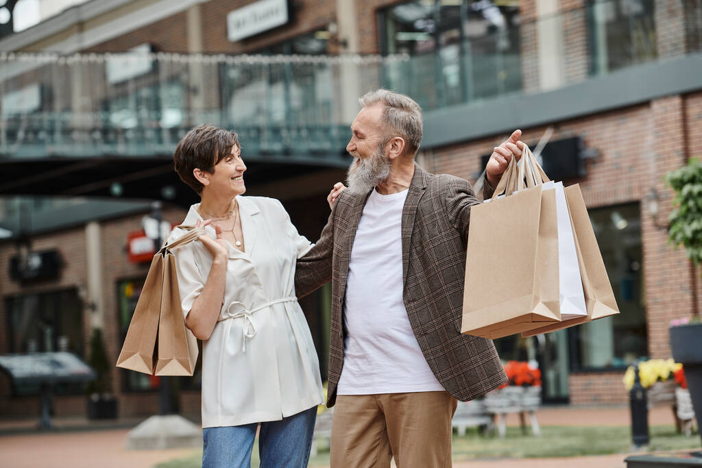 счастливая пара с сумками, пожилой мужчина и женщина смотрят друг на друга, открытый торговый центр - Фото, изображение
