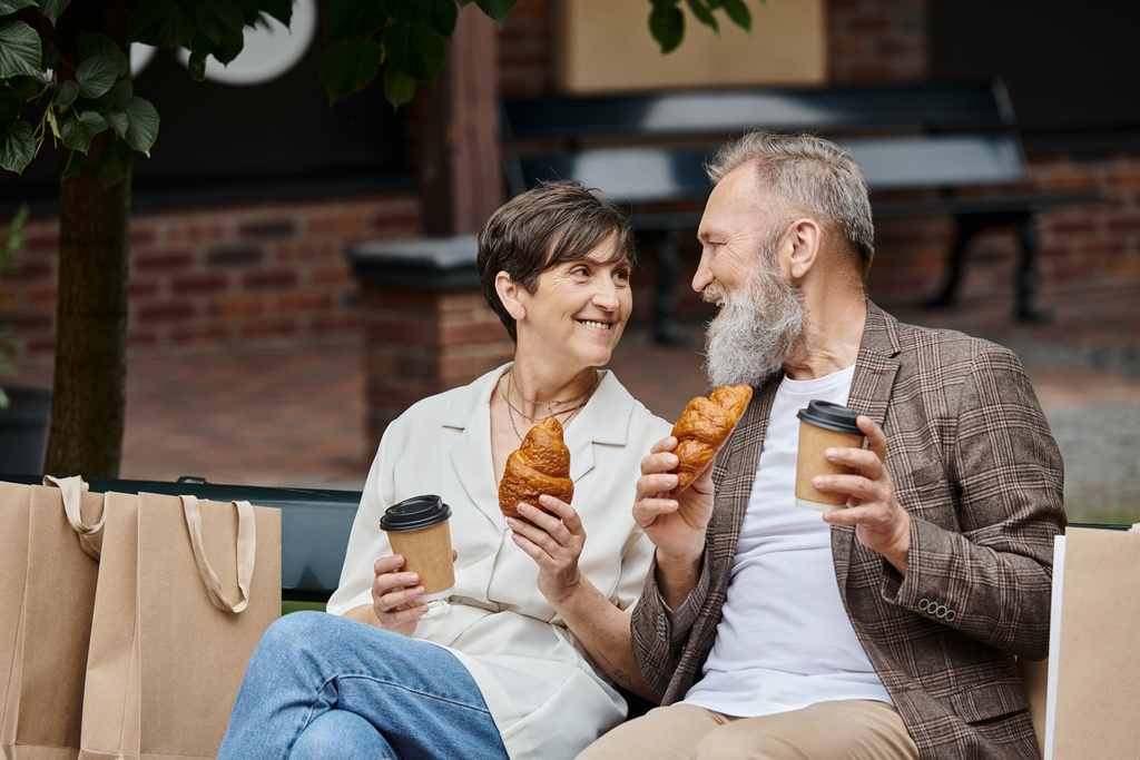 heureux couple âgé tenant croissants et café à emporter, gobelets en papier, sacs à provisions, sortie, urbain - Photo, image