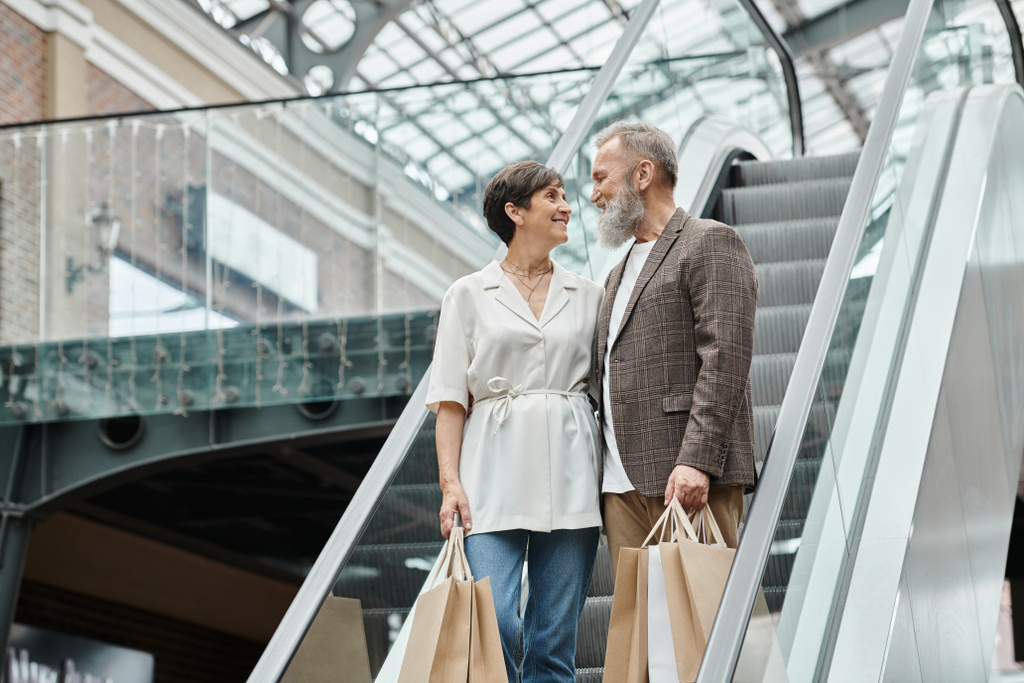 homme et femme âgés positifs debout sur l'escalator, sacs à provisions, se regardant dans le centre commercial - Photo, image