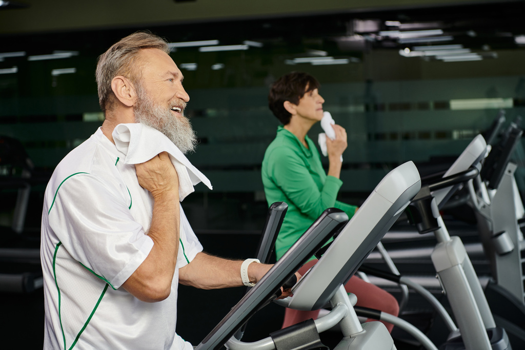 hombre mayor y barbudo limpiando el sudor con toalla, mujer borrosa en el fondo, haciendo ejercicio en el gimnasio - Foto, Imagen