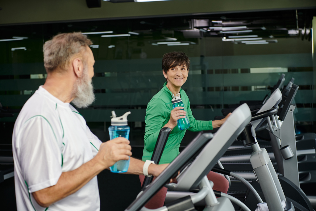 radosna kobieta patrząc na starszego mężczyznę, męża i żonę ćwiczących na siłowni, trzymając butelki sportowe - Zdjęcie, obraz