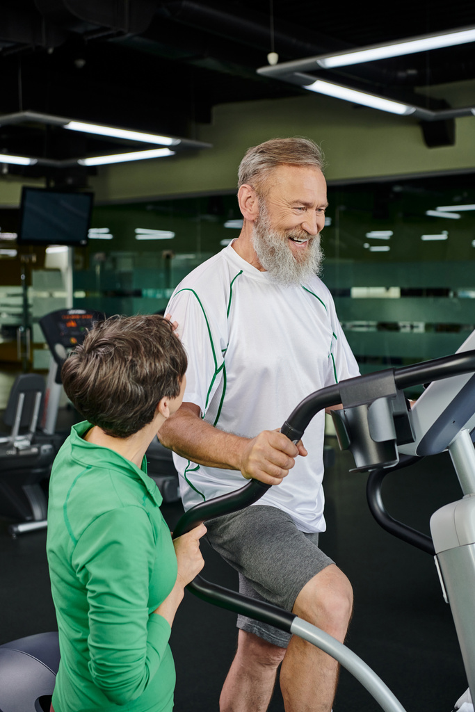 homme âgé heureux et barbu faisant de l'exercice près d'une femme en salle de gym, mari et femme, personnes âgées actives, sport - Photo, image