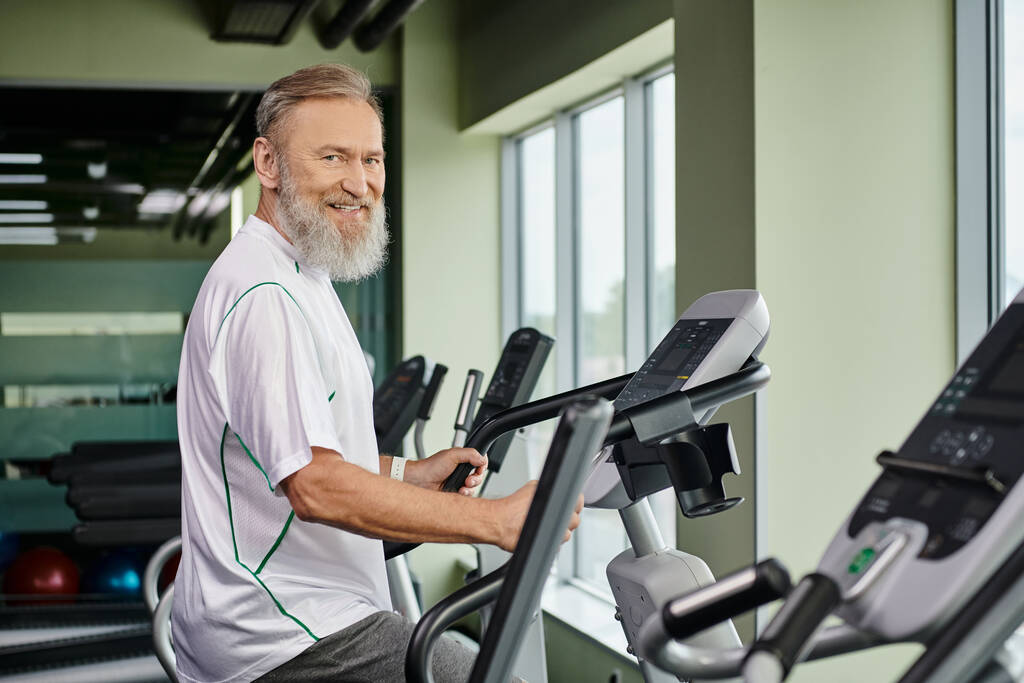 χαρούμενος γενειοφόρος άνθρωπος που εργάζονται για το μηχάνημα άσκησης, ηλικιωμένοι στο γυμναστήριο, ενεργός ανώτερος, γυμναστήριο και τον αθλητισμό - Φωτογραφία, εικόνα