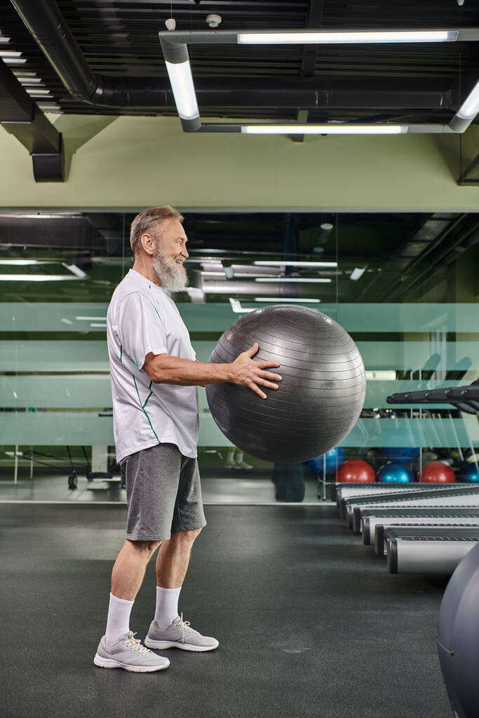 alegre anciano sosteniendo pelota de fitness en el gimnasio, ejercicio, fitness y deporte, estilo de vida activo - Foto, imagen