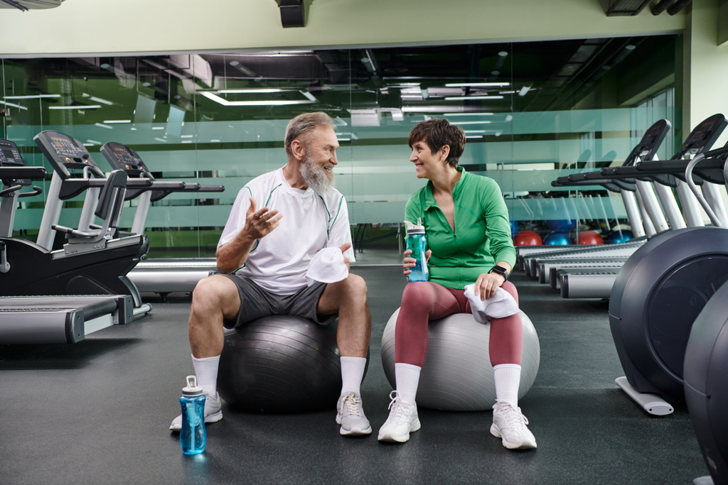 deportivo pareja de ancianos, alegre hombre y mujer sentado en pelotas de fitness, sosteniendo botellas con agua - Foto, imagen