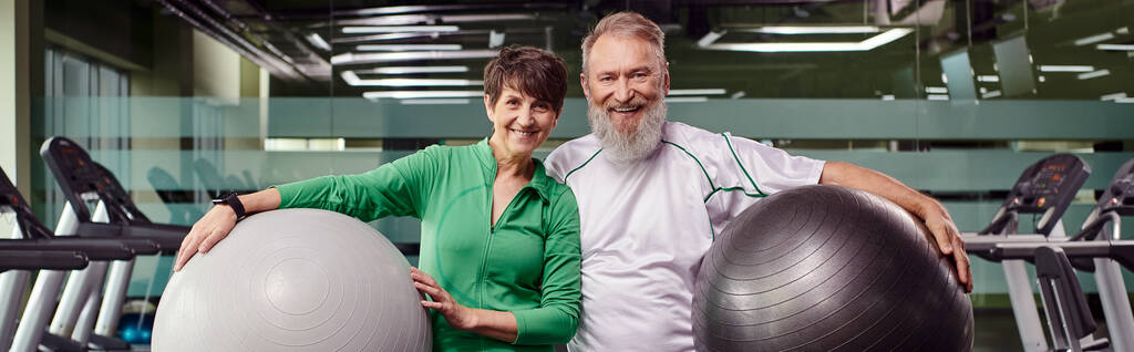 σπορ ηλικιωμένο ζευγάρι, χαρούμενος άνδρας και γυναίκα κρατώντας μπάλες γυμναστικής, ενεργοί ηλικιωμένοι στο γυμναστήριο, πανό - Φωτογραφία, εικόνα