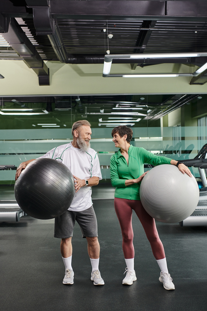 starší pár, radostný muž a žena držící fitness míče, aktivní senioři dívající se jeden na druhého - Fotografie, Obrázek