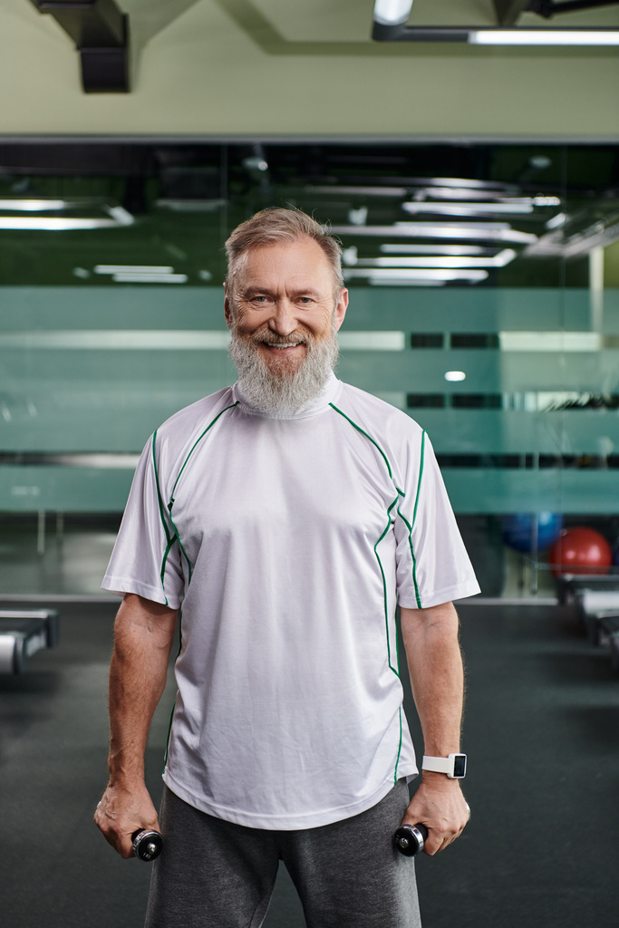 счастливый и бородатый пожилой мужчина улыбается и смотрит в камеру, тренируется с гантелями в тренажерном зале - Фото, изображение