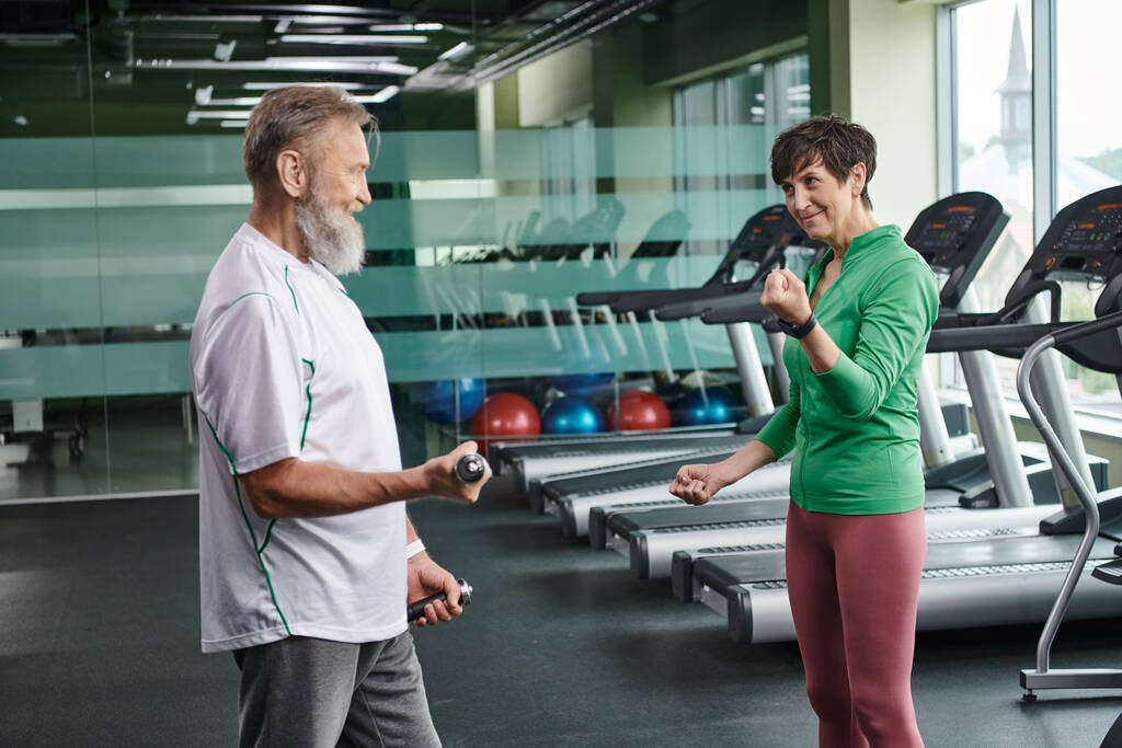 Mutlu kadın sakallı adama bakıyor spor salonunda dambıllarla çalışıyor, aktif yaşlılar, yaşam tarzı... - Fotoğraf, Görsel