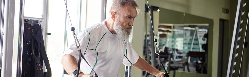 hombre anciano motivado con barba haciendo ejercicio en la máquina de ejercicio en el gimnasio, atleta, activo, bandera - Foto, imagen