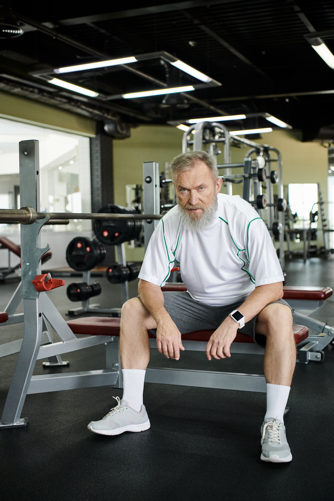 Müder älterer Mann mit Bart blickt nach dem Training in die Kamera, sitzt im Fitnessstudio auf einem Trainingsgerät - Foto, Bild