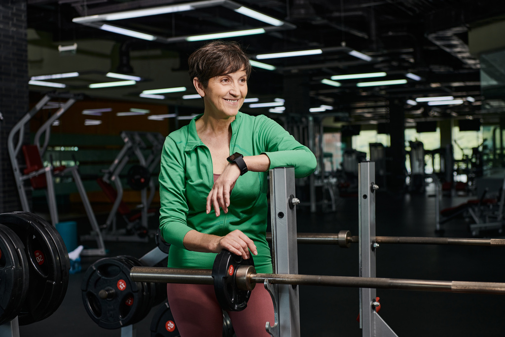 heureuse femme âgée souriante et debout près de l'haltère, plaques de poids, séance d'entraînement en salle de gym, motivation - Photo, image