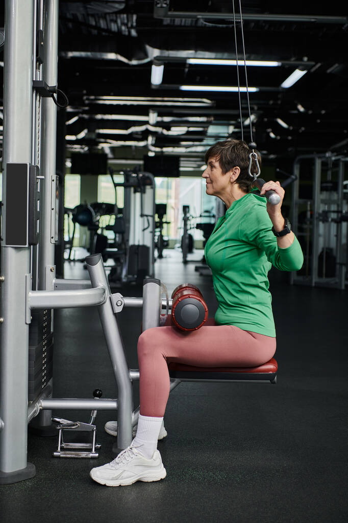silná a motivovaná starší žena cvičící v tělocvičně, zralé fitness, cvičební stroj, boční pohled - Fotografie, Obrázek