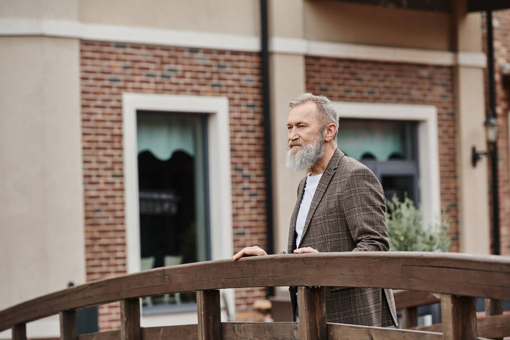 bebaarde oudere man met grijs haar op houten brug, wegkijkend, denkend, stedelijk decor - Foto, afbeelding