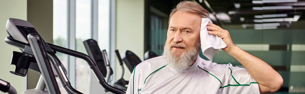 horizontal, pancarta, hombre mayor con barba limpiando el sudor con toalla después de hacer ejercicio en el gimnasio, activo - Foto, Imagen