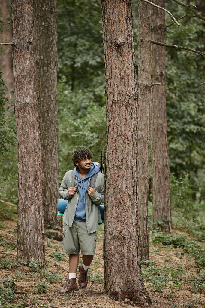 χαμογελαστός νεαρός ινδός ταξιδιώτης με σακίδιο πλάτης στέκεται κοντά σε δέντρο ενώ ξεκουράζεται στο δάσος - Φωτογραφία, εικόνα