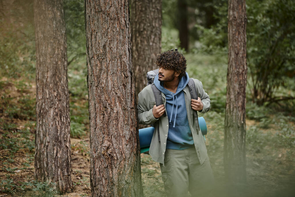 кудрявый молодой индийский путешественник с рюкзаком, стоящим возле дерева в лесу - Фото, изображение