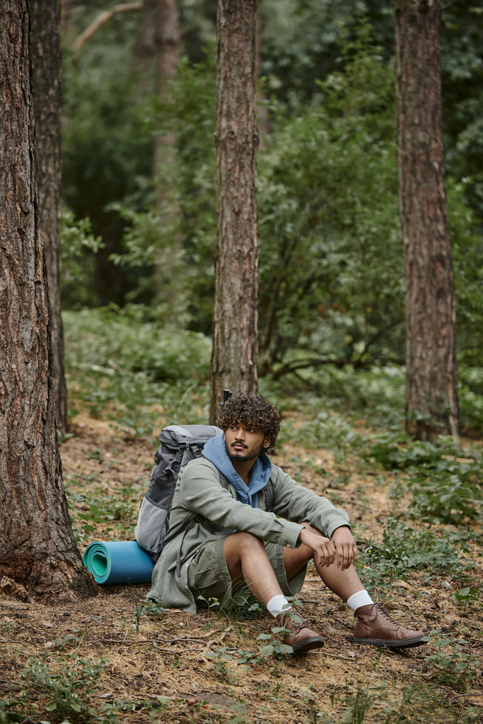 кучерявий молодий індійський пішохід з рюкзаком і похідними полюсами відпочиває, сидячи біля дерев у лісі - Фото, зображення