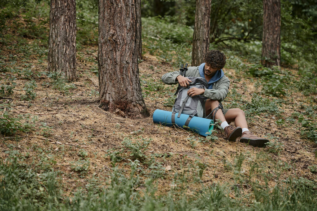 Kıvırcık genç Hintli gezgin ormanda dinlenirken sırt çantasını açıyor. - Fotoğraf, Görsel