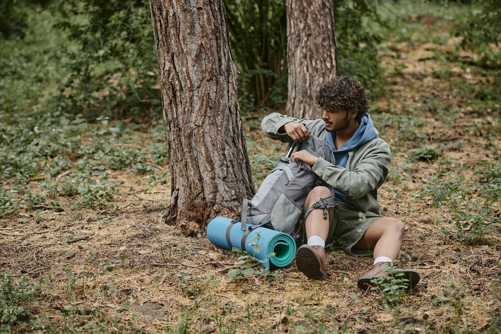 Genç Hintli yürüyüşçü ormanda yerde dinlenirken spor minderinin yanında sırt çantasını açıyor. - Fotoğraf, Görsel