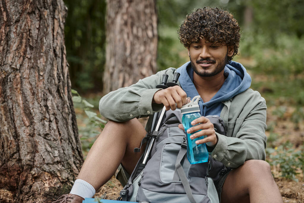 Lächelnder junger indischer Wanderer mit Sportflasche in der Nähe von Rucksack, während er sich im Wald ausruht - Foto, Bild