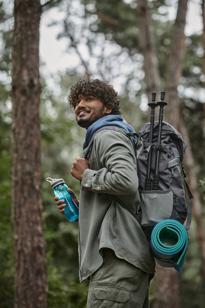 позитивний молодий індійський мандрівник тримає рюкзак і спортивну пляшку під час прогулянки в лісі - Фото, зображення