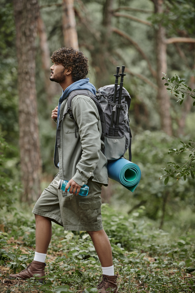 vue latérale du jeune touriste indien avec sac à dos et bouteille de sport marchant en forêt - Photo, image
