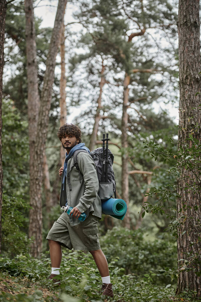 kudrnatý indický cestovatel drží batoh a sportovní láhev při chůzi v lese - Fotografie, Obrázek