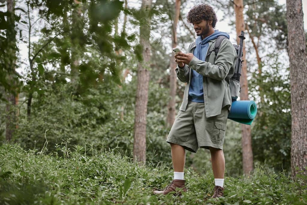 smiling young indian tourist with backpack using smartphone while standing in forest - Photo, Image