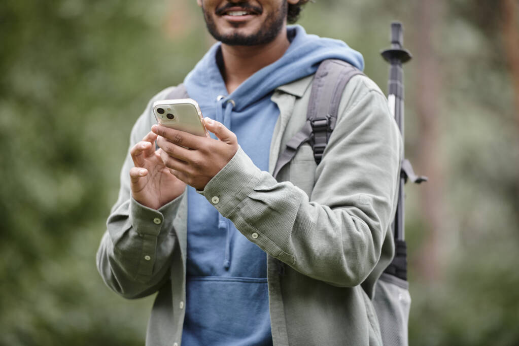 vista ritagliata di escursionista indiano sorridente con zaino utilizzando smartphone nella foresta sfocata - Foto, immagini