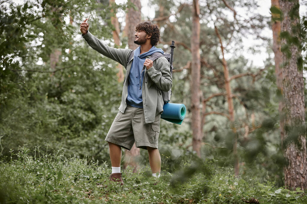 усміхнений індійський пішохід з рюкзаком бере селфі на смартфон у розмитому лісі - Фото, зображення
