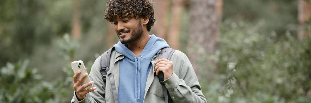 pozitivní mladý indický turista s batohem pomocí smartphonu v lese, banner - Fotografie, Obrázek