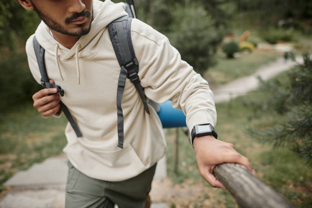 bijgesneden uitzicht op jonge indiaanse toerist met rugzak en smartwatch wandelen in de buurt van hek in het bos - Foto, afbeelding