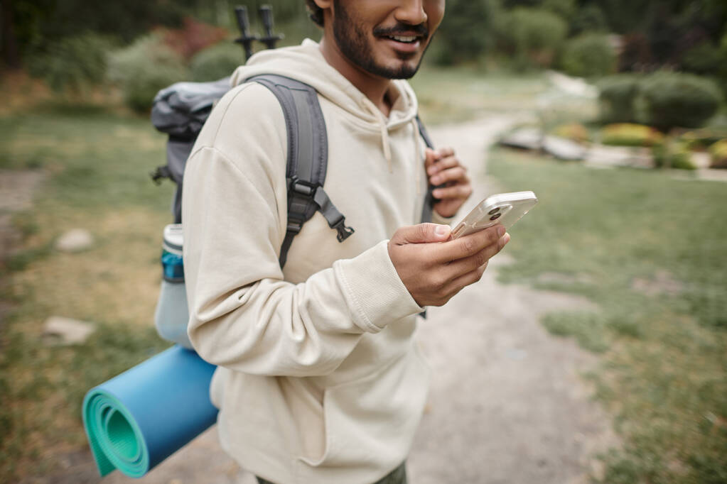 abgeschnittene Ansicht eines lächelnden indischen Reisenden mit Rucksack und Smartphone im Wald - Foto, Bild