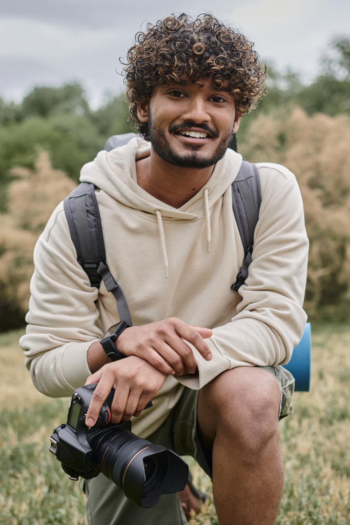 gioioso fotografo indiano con fotocamera professionale e guardando la fotocamera nella foresta, verticale - Foto, immagini