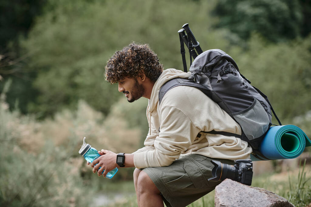 šťastný indický turista sedí s batohem a drží láhev s vodou v lese, dobrodružství - Fotografie, Obrázek