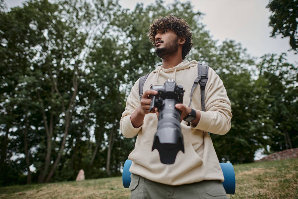 homem indiano concentrado segurando câmera digital e de pé com mochila na floresta, fotógrafo - Foto, Imagem