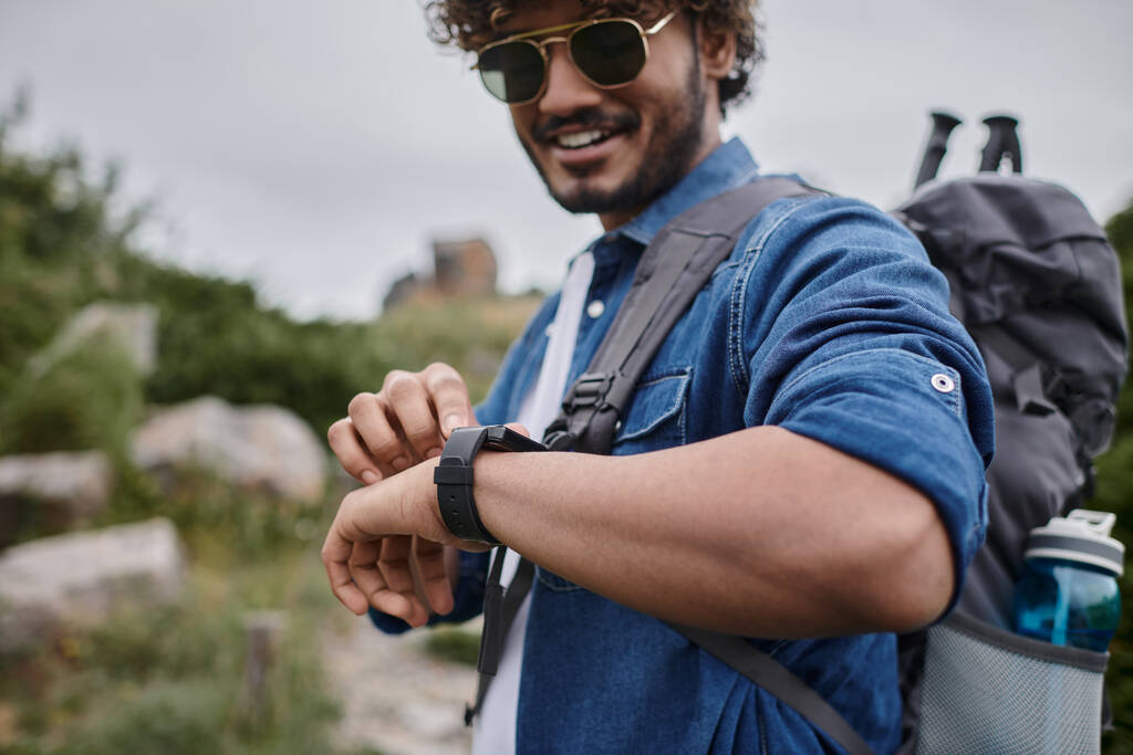 счастливый индиец в солнечных очках и джинсовой рубашке время проверки на наручные часы, турист с дорожным снаряжением - Фото, изображение
