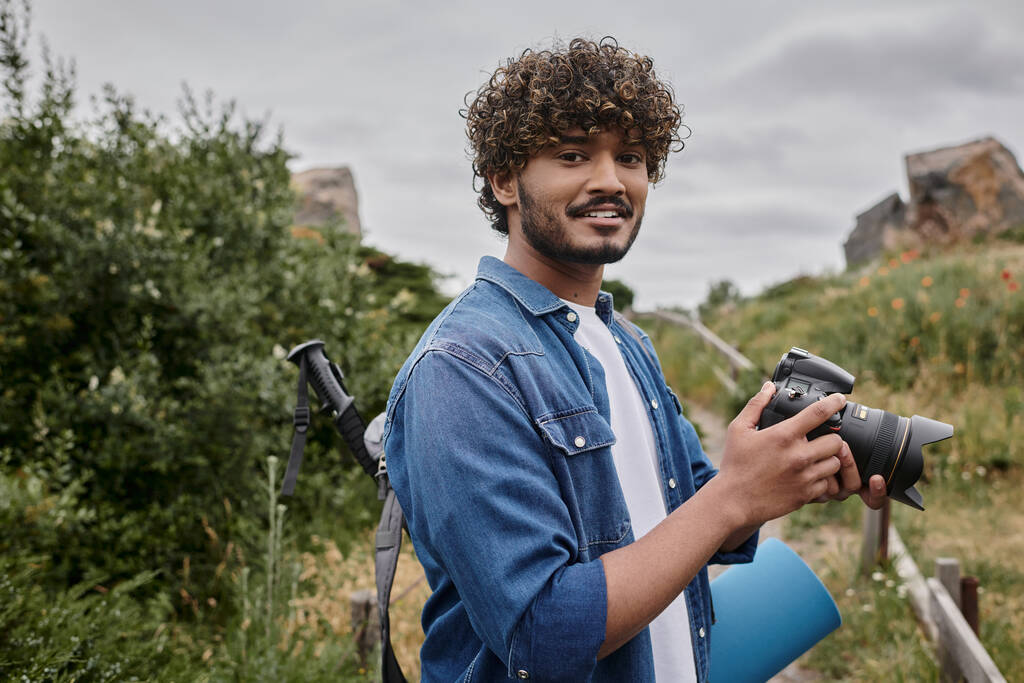concepto de viaje y fotografía, mochilero indio rizado que sostiene la cámara digital durante el viaje por la naturaleza - Foto, Imagen