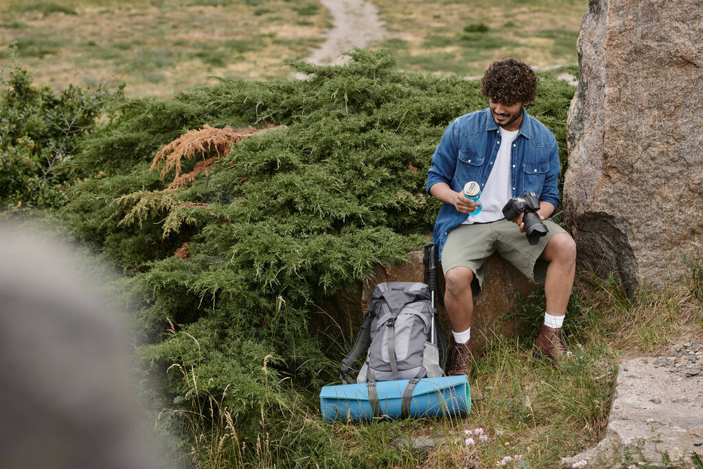Turist ve doğa kavramı, Hintli adam kayanın üzerinde otururken su ve kamerayla şişeyi tutuyor. - Fotoğraf, Görsel