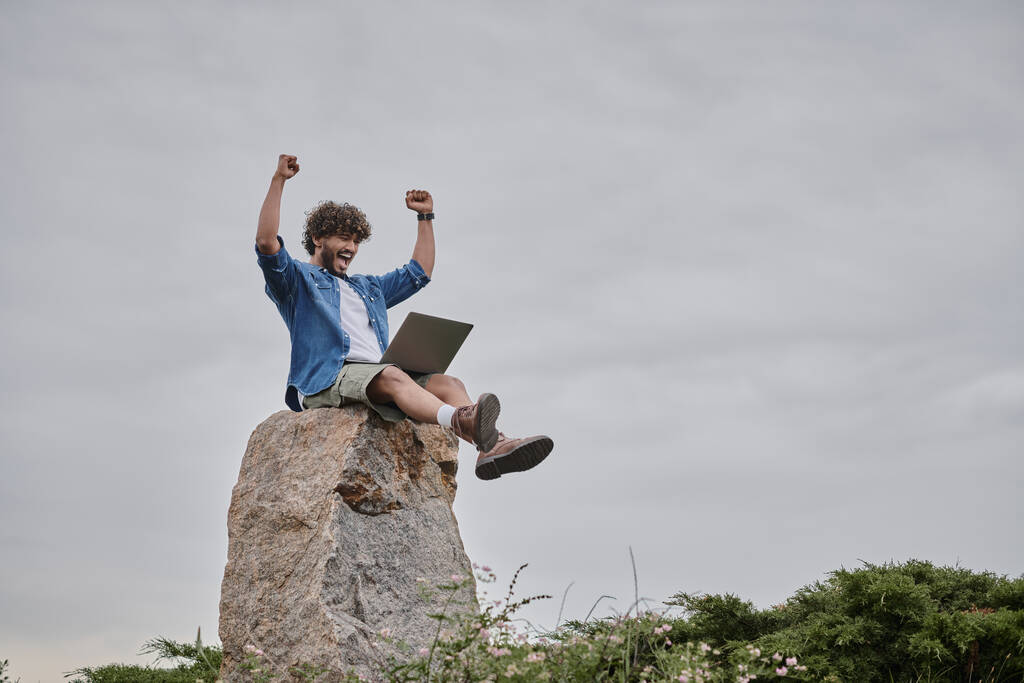 digital nomad concept, excited indian freelancer celebrating win while using laptop, sitting on rock - Photo, Image
